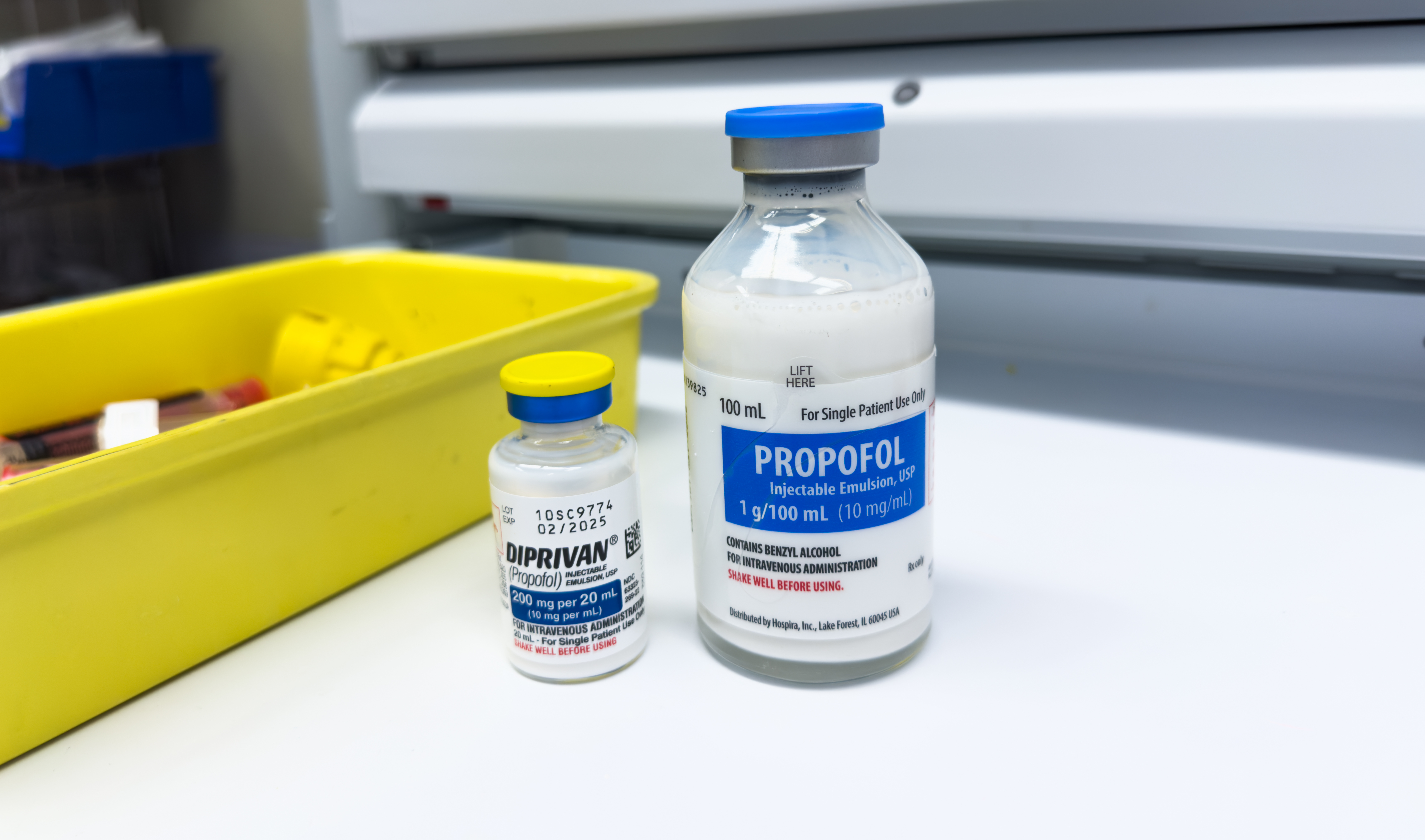 Two vials of propofol, a milky liquid, rest on a hospital shelf next to a small yelllow bin 