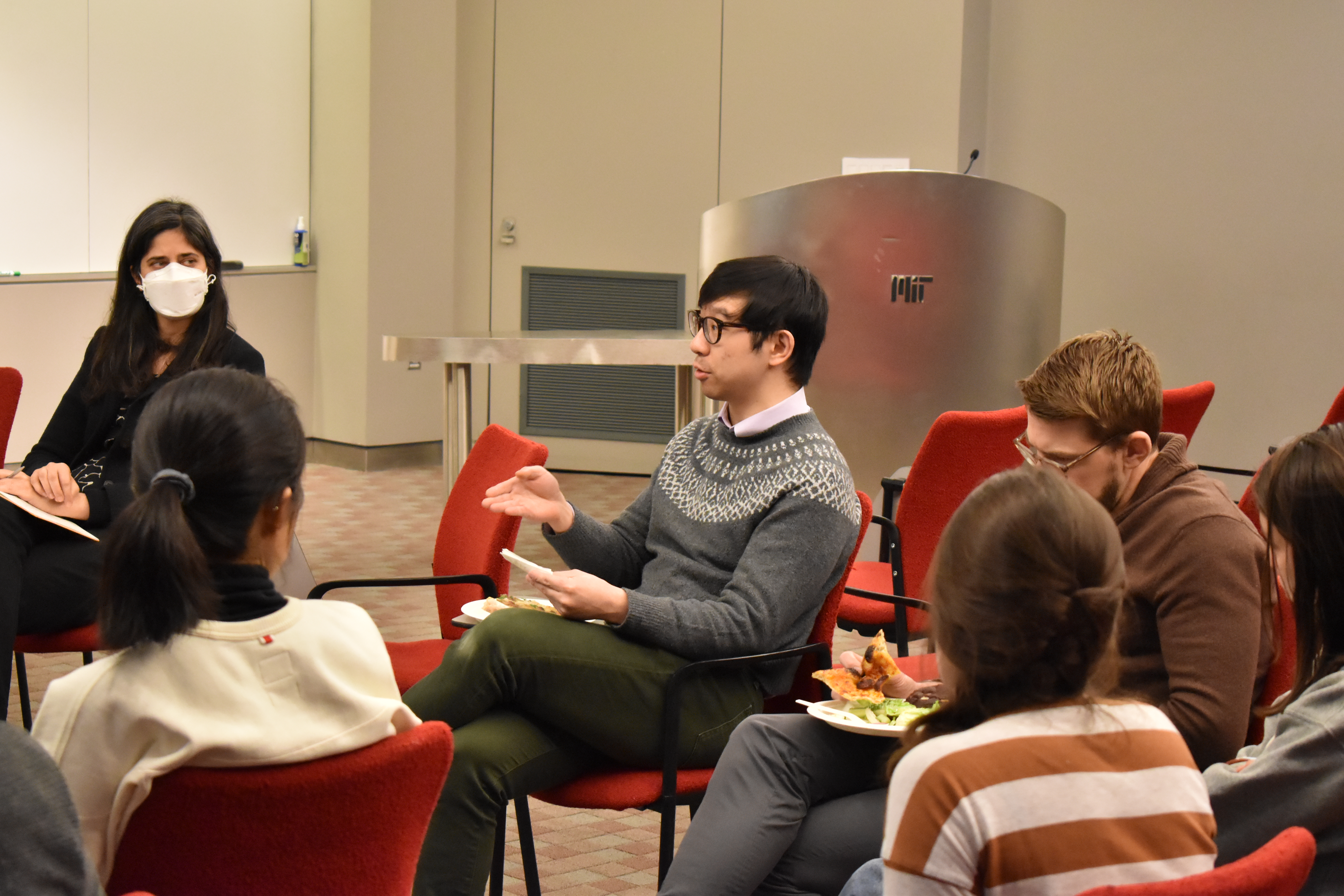 A man sitting in a chair gestures as he looks to the left out of the frame to the individual whom he is addressing. Other people are seated nearby and appear to be listening.