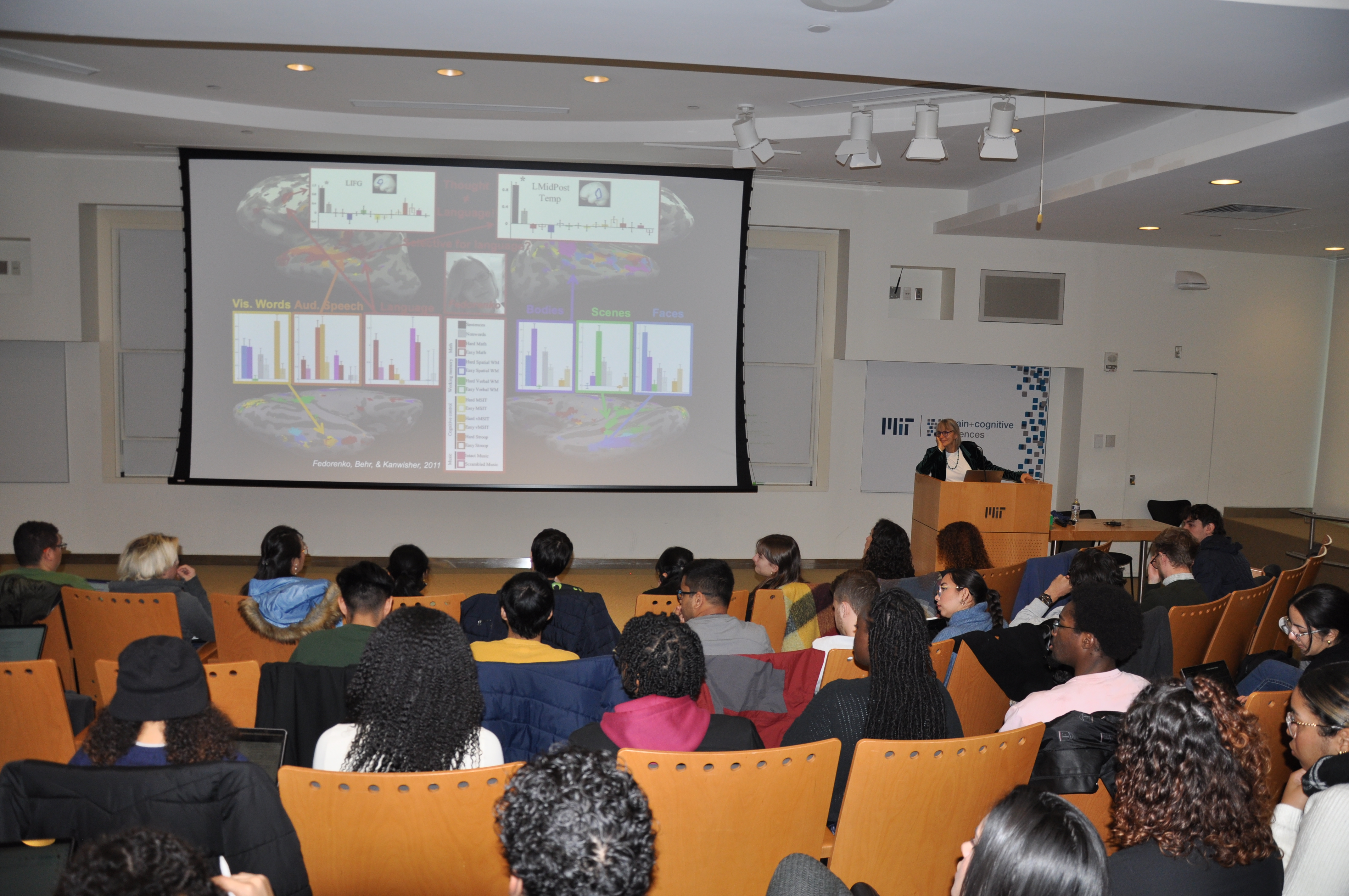 Nancy Kanwisher stands at the lectern in a large theater style lecture hall. A full audience is facing her and the screen.