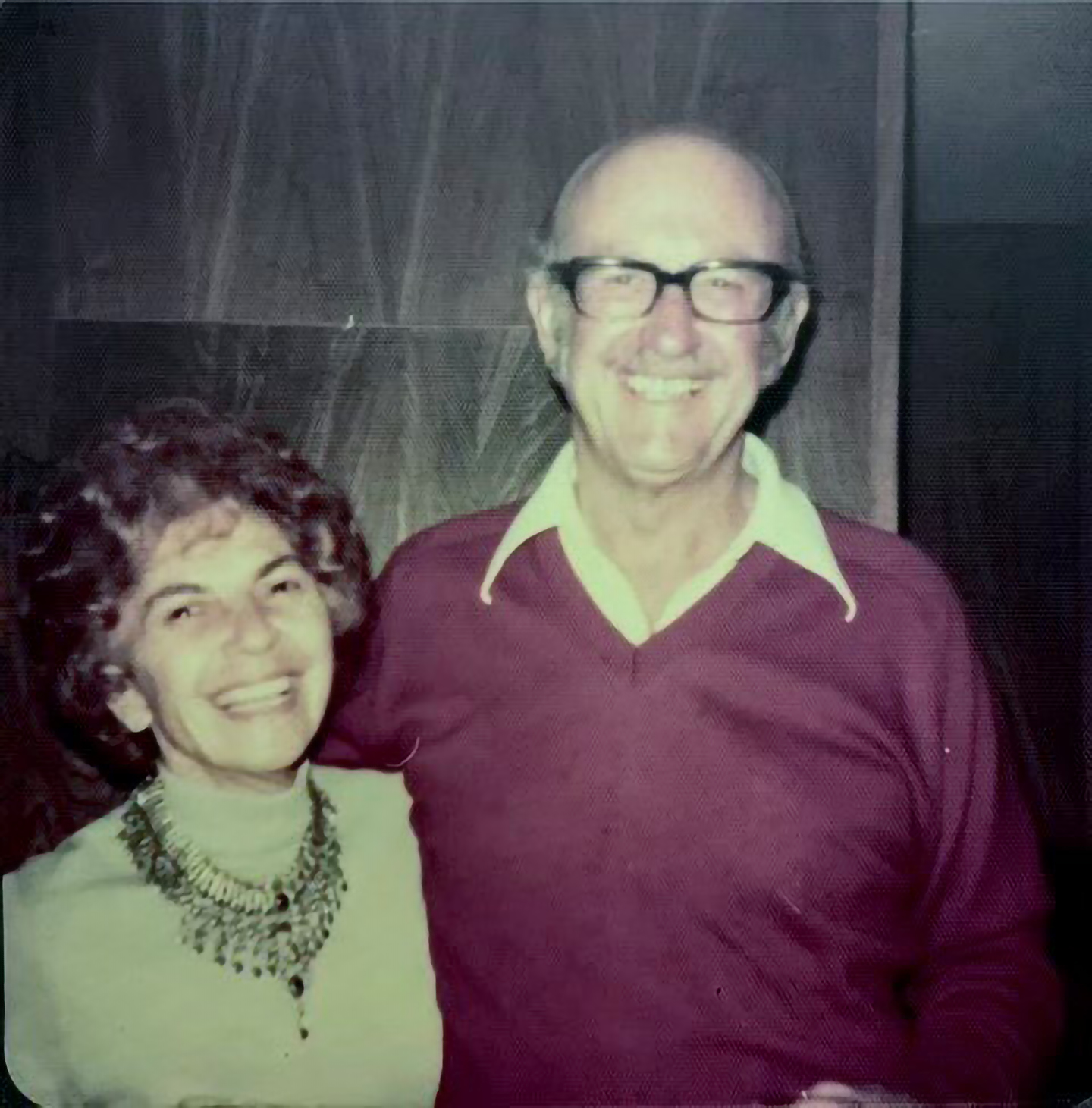 A picture of Betty and Dana Fisher standing together and smiling in front of a wood paneled wall.