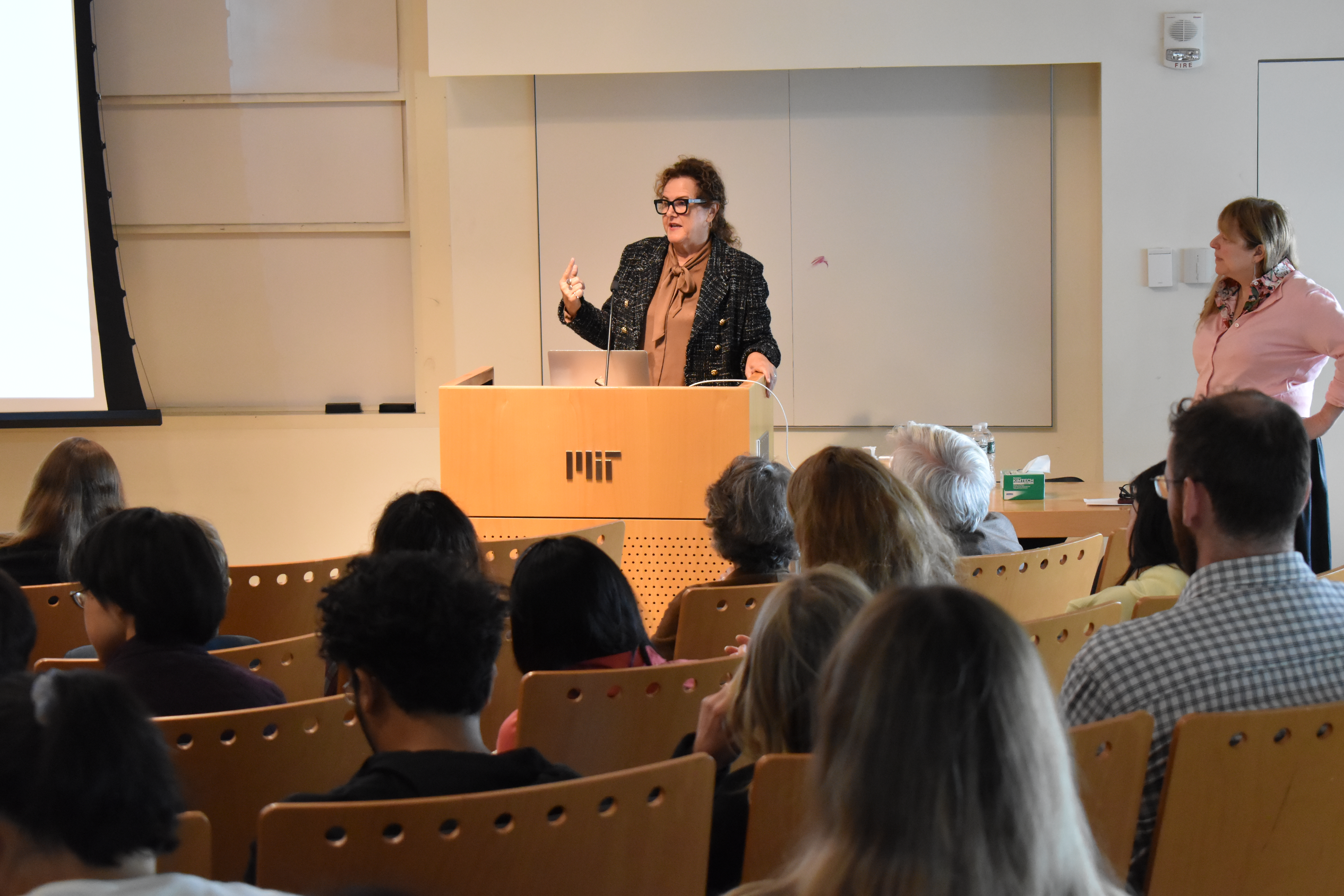 Erin Schuman stands and gestures behnind a wooden lectern with the MIT logo on it. Her audience is in the foreground sitting in rows of auditorium style chairs.