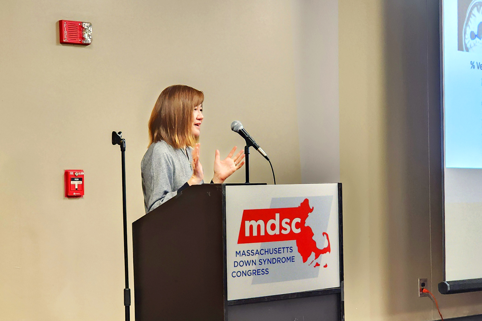 Li-Huei Tsai smiles and gestures as she stands at a podium with a placard displaying the Massachusetts Down Syndrome Congress logo