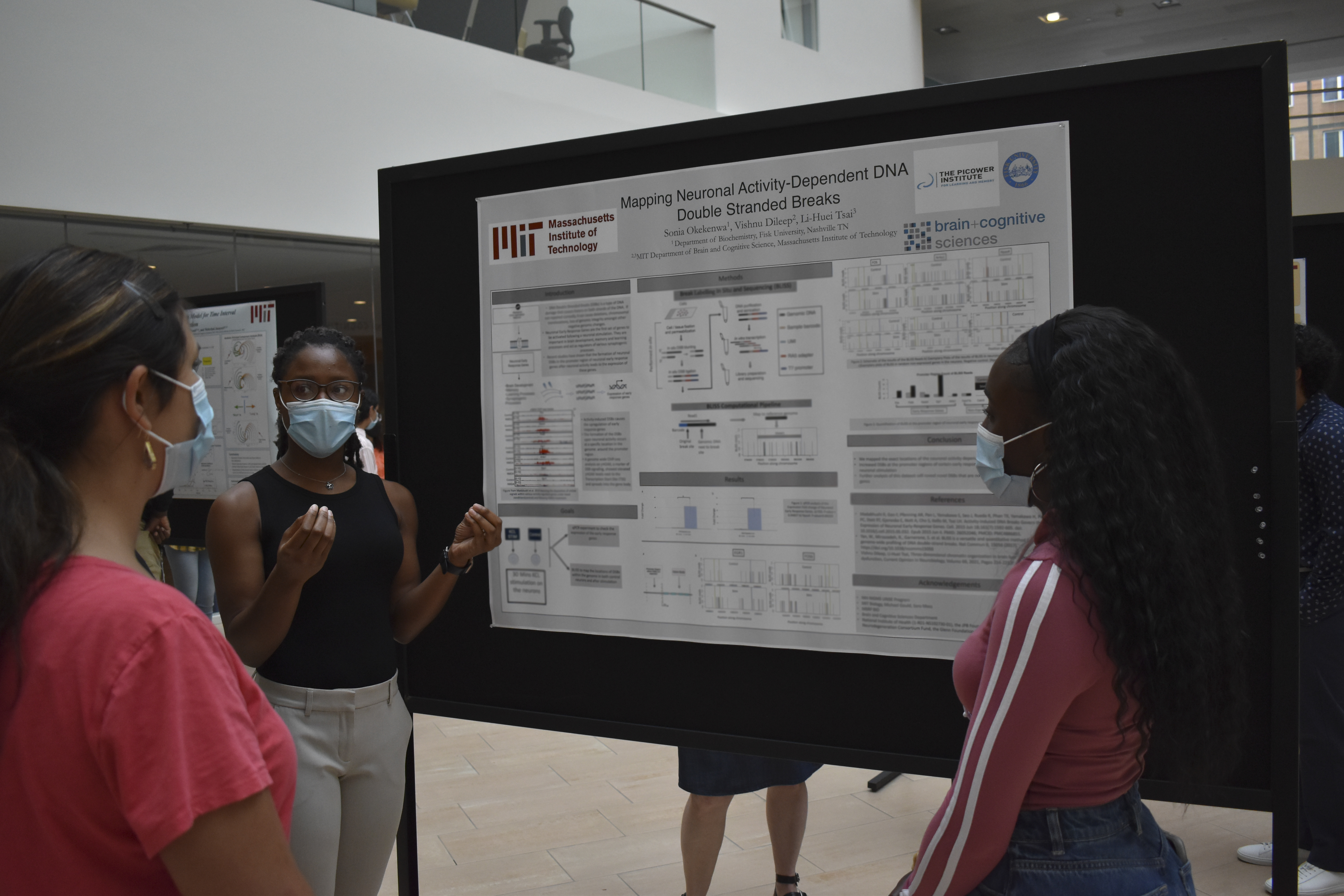 A woman stands in front of a big poster on a black stand and gestures as she explains the contents to two other women looking on.