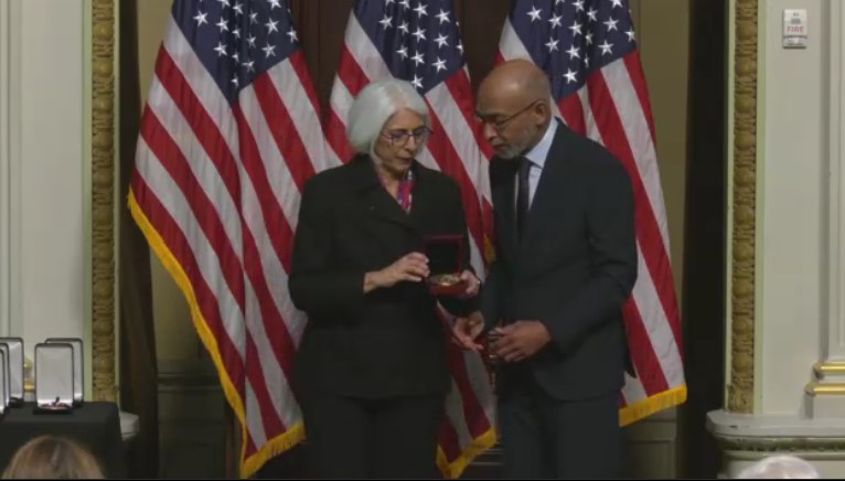 Emery Brown is handed a medal by a woman as they stand before a backdrop of three American flags in an ornate room of the White House.