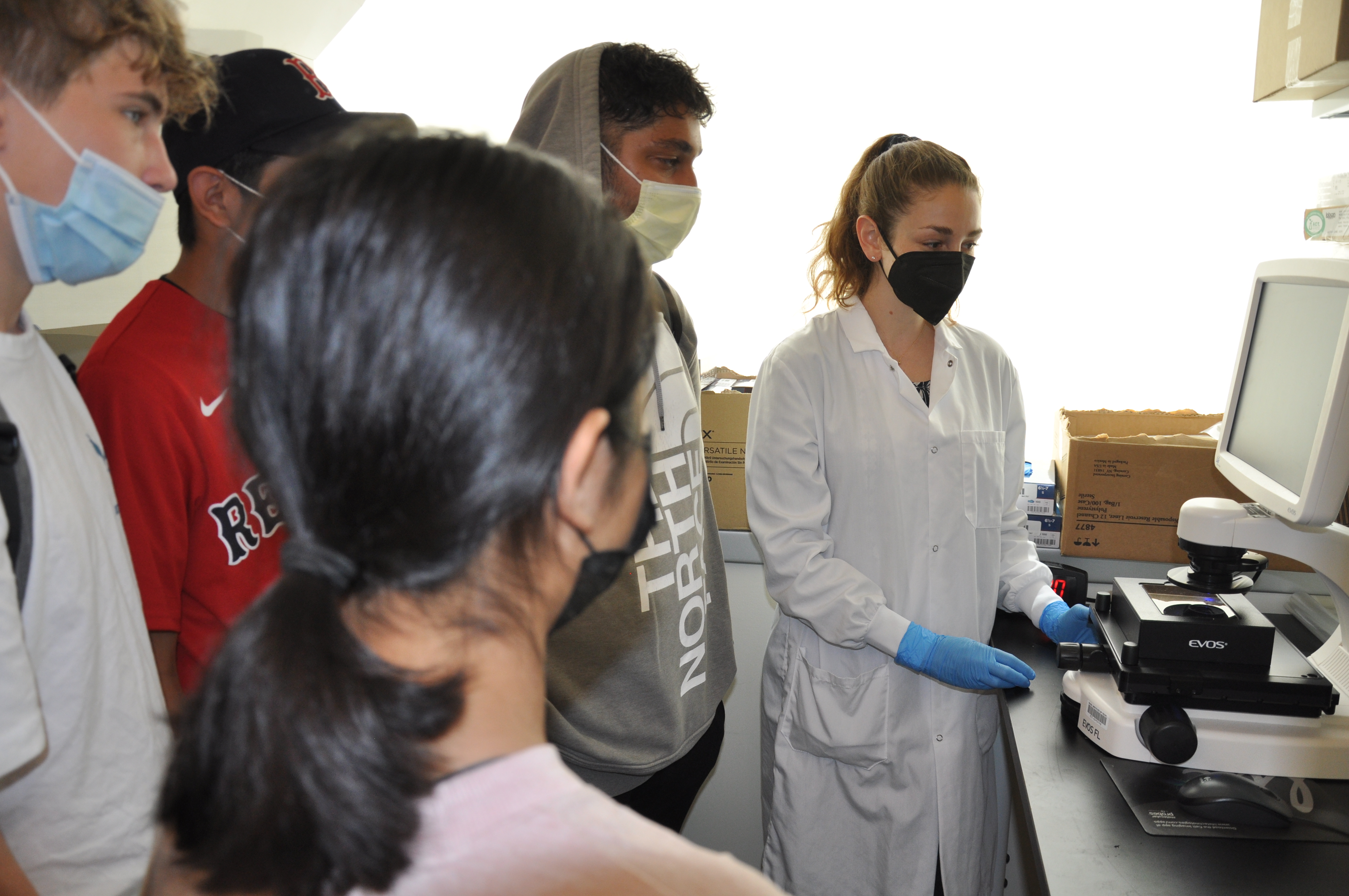 A woman in a white lab coat works at a microscope as a group of students watches