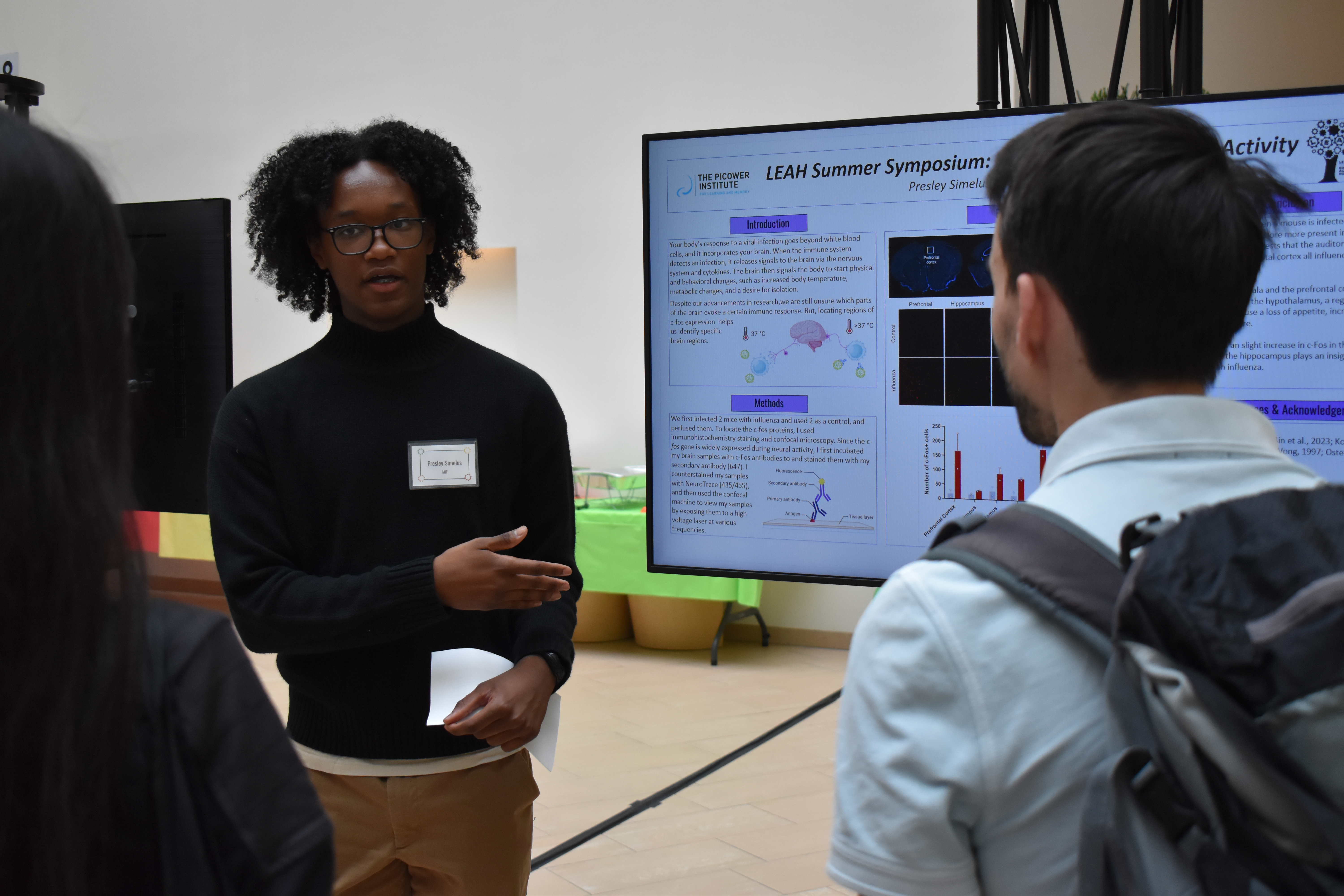 Presley Simelus stands at a large electronic display showing his research poster as two people stand in the foreground listening to him speak.