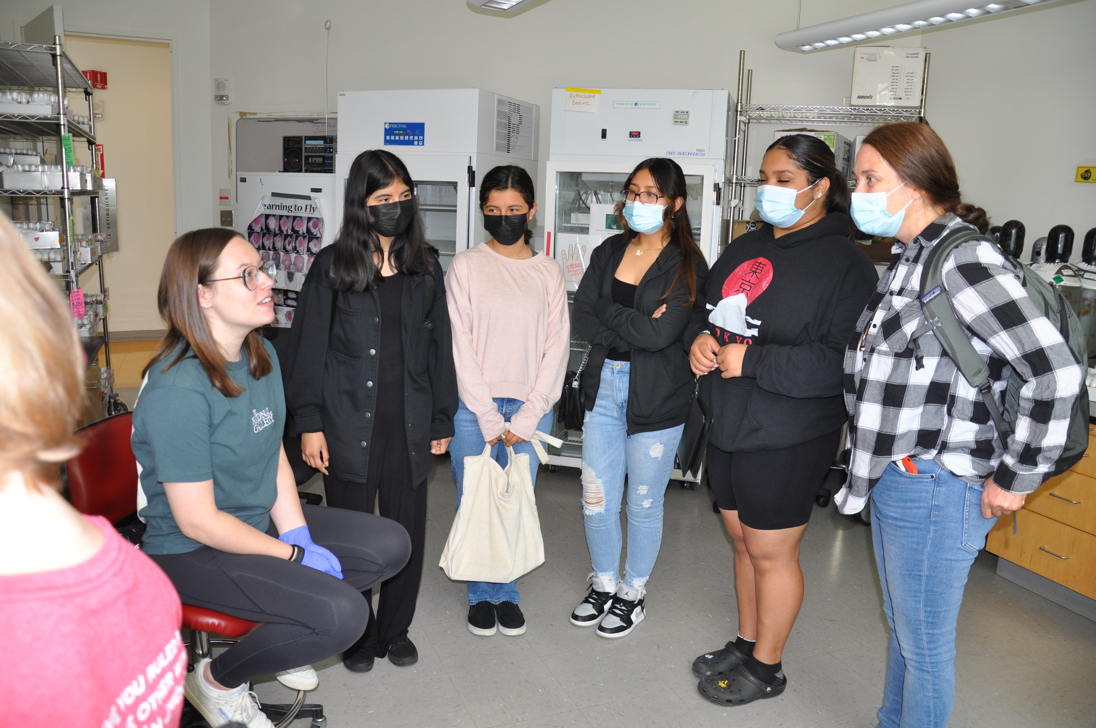 A woman sits in a chair in a lab and talks to a group of students and their teacher standing in a semicircle around her