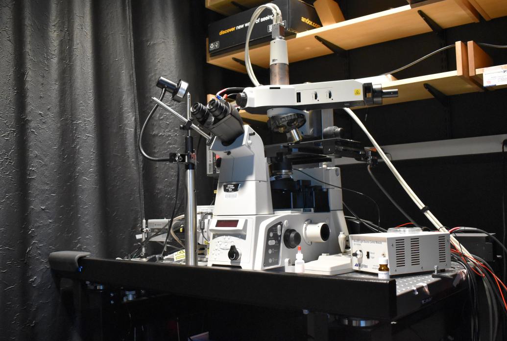 A white microscope with many wires and attachments sits on a black table