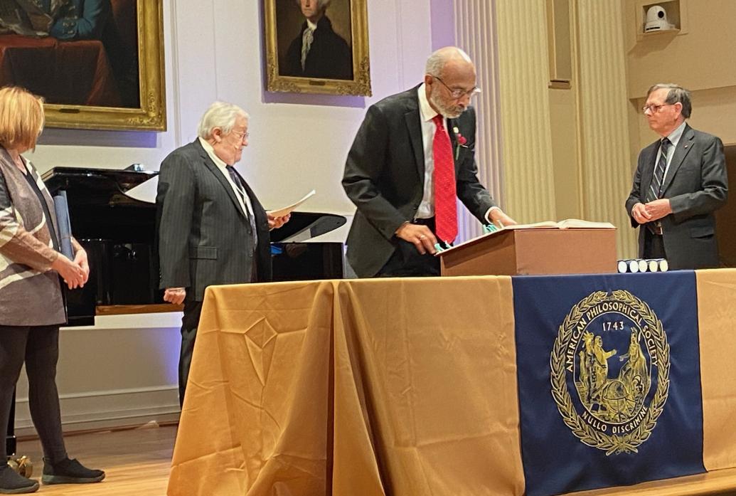 Emery Brown examines a book on a lectern in an ornate room with classical american portraits on the wall. The table on which the lectern rests is draped with a golden cloth and the blue seal of the American Philosophical Society.