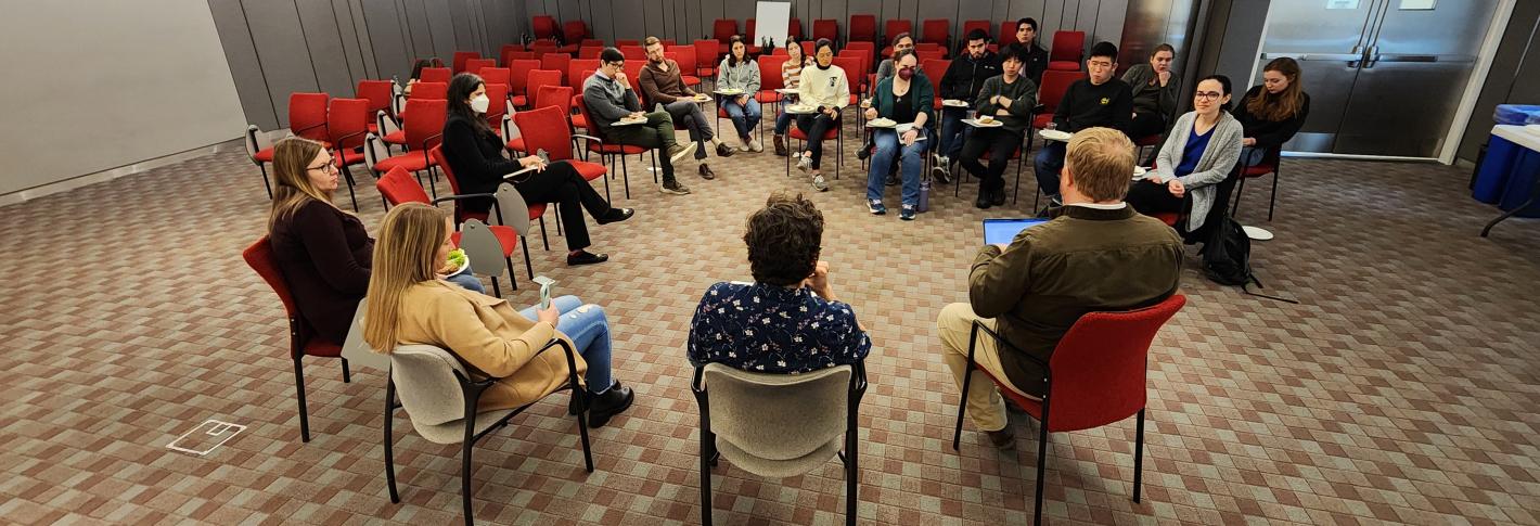 A group of about 16 people having a discussion sits arranged in a circle in red chairs in a large, bright room. The people have serious expressions on their faces. though a few are taking bites of pizza. In the background are open pizza boxes.