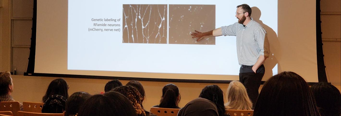 Brady Weissbourd stands at the front of an auditorium with images describing an experiment on the screen and an audience of teenagers sitting in the audience