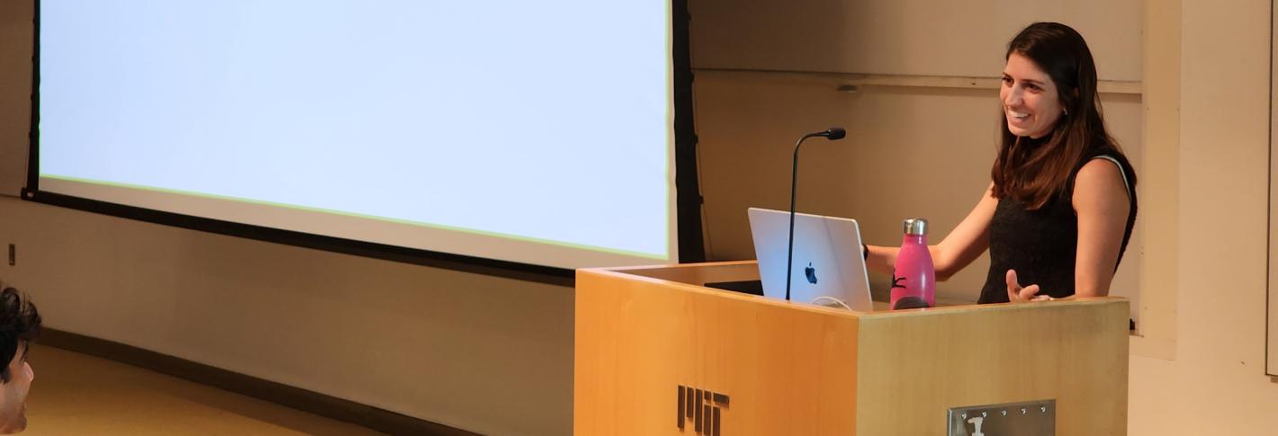 Gabi Drummond stands at a podium and smiles at the audience as she defends her thesis. The title on the screen is "The role of locus coeruleus norepinephrine in reinforcement learning"