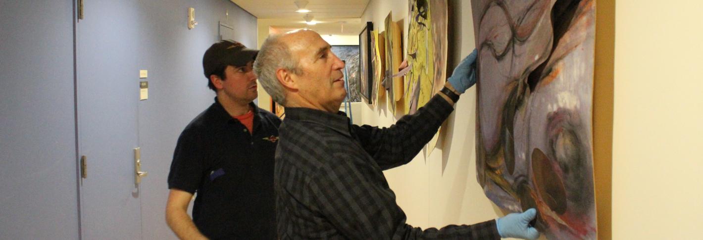 Todd Siler holds a painting up against a wall while an installer helps him assess its placement