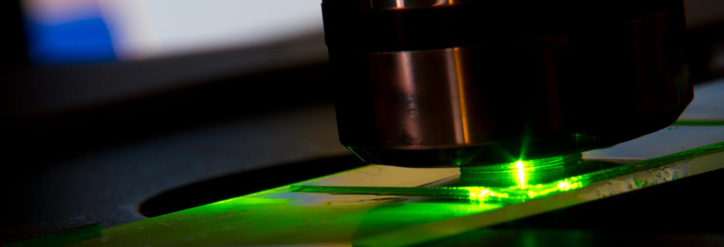 A closeup of a microscope stage. The round metal objective shines a bright green laser light