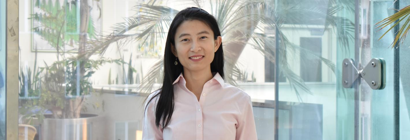 Linlin Fan smiles as she stands in a glassy room with reflections of large tropical plants in the windows behind her.