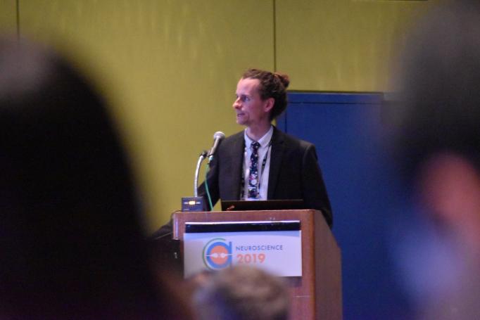 Peter Finnie stands at a podium, seen between the blurred heads of two audience members